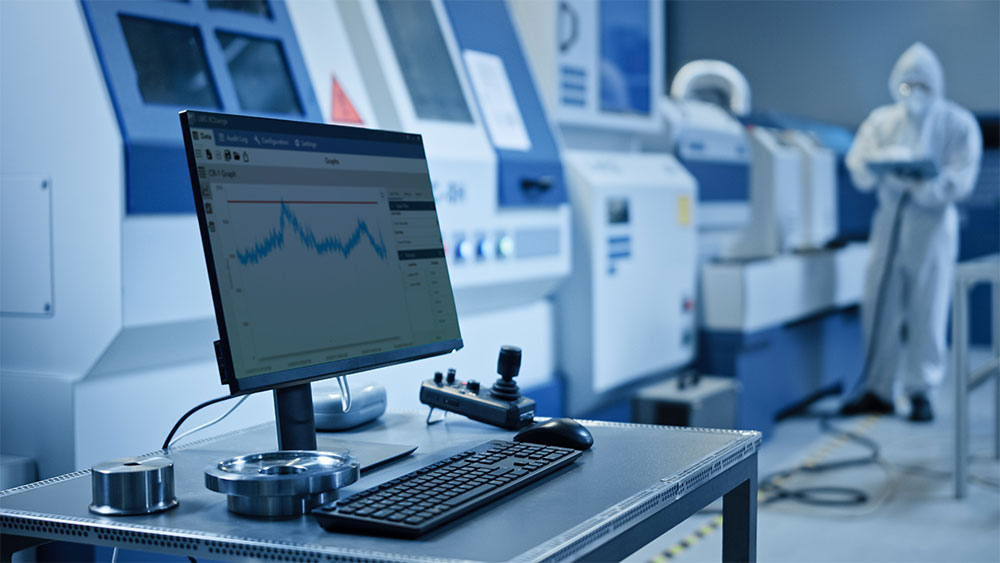 photo d'un ordinateur portable, d'un clavier et d'une souris posés sur un bureau dans une salle blanche avec un technicien de salle blanche effectuant un travail debout