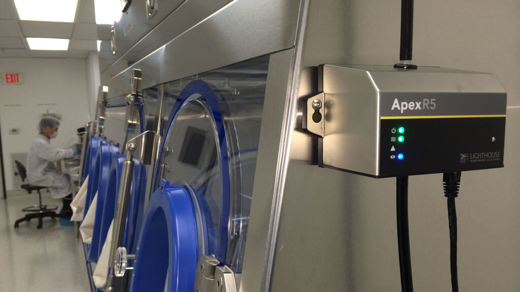 photo du produit ApexR5 monté sur un mur dans une salle blanche avec un technicien de salle blanche portant un masque effectuant un travail à un bureau